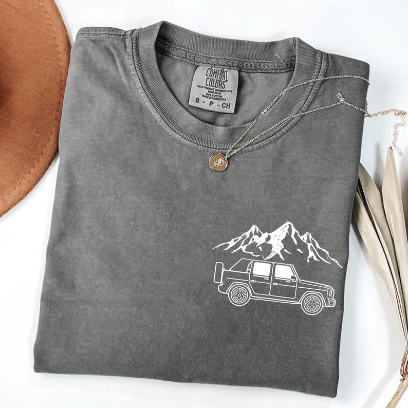 May include: A charcoal gray t-shirt with a white mountain and truck graphic. The shirt has a crew neck and short sleeves. A silver necklace with a pendant is draped across the shirt. The shirt is labeled "Comfort Colors".