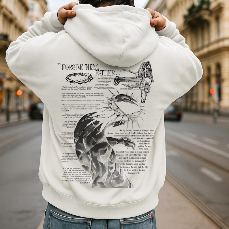 May include: White hoodie with a graphic design on the back. The design includes text that reads "FORGIVE THEM, FATHER..." along with illustrations of a crown of thorns, a cross, and other religious imagery. The hoodie is a casual garment.