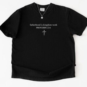 以下が含まれることがあります： fatherhood is kingdom workおよびPROVERBS 22:6という白いテキストが、小さな白い十字架のグラフィックの上に中央揃えで配置された黒い半袖Tシャツ。小さなペンダントが付いた銀色のチェーンネックレスが襟元に置かれています。