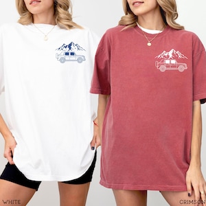 May include: Two oversized t-shirts, one white and one crimson, each featuring a mountain and vehicle graphic on the chest. The white shirt has a navy blue design, while the crimson shirt has a white design. The shirts are casual wear.