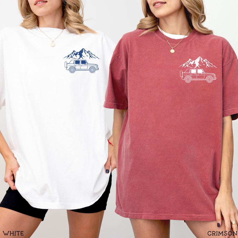 May include: Two oversized t-shirts, one white and one crimson, each featuring a mountain and vehicle graphic on the chest. The white shirt has a navy blue design, while the crimson shirt has a white design. The shirts are casual wear.