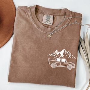 May include: A brown Comfort Colors t-shirt featuring a white graphic of a mountain range and a truck. The shirt has a crew neck and short sleeves. A silver necklace with a pendant is also visible.