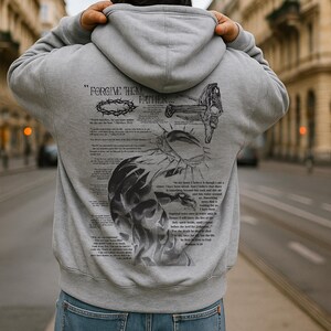 May include: Gray hoodie with a hood and a large graphic print on the back. The print includes the text "FORGIVE THEM FATHER" along with illustrations of a crown of thorns, a cross, and scriptural text. The hoodie is made of a soft material.