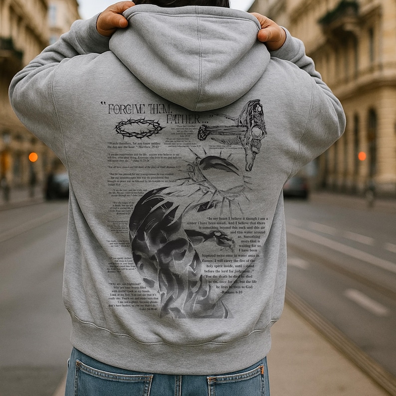 May include: Gray hoodie with a hood and a large graphic print on the back. The print includes the text "FORGIVE THEM FATHER" along with illustrations of a crown of thorns, a cross, and scriptural text. The hoodie is made of a soft material.