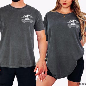 May include: Two dark gray t-shirts with a white mountain and off-road vehicle graphic. The shirts have a crew neck and short sleeves. One person is wearing black shorts. The other person is wearing black shorts.