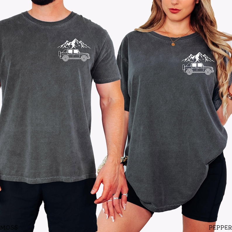 May include: Two dark gray t-shirts with a white mountain and off-road vehicle graphic. The shirts have a crew neck and short sleeves. One person is wearing black shorts. The other person is wearing black shorts.