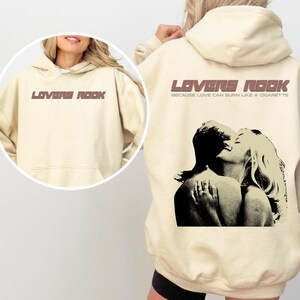 May include: A cream-colored hoodie with a graphic print of a couple embracing. The text "LOVERS ROCK" is printed in brown on the front of the hoodie, and the text "BECAUSE LOVE CAN BURN LIKE A CIGARETTE" is printed in brown below the graphic.