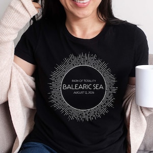 May include: Black t-shirt featuring a white circular design with radiating lines and the text "PATH OF TOTALITY, BALEARIC SEA, AUGUST 12, 2026." The shirt is a crew neck style.