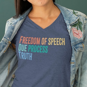 May include: Navy blue V-neck t-shirt featuring the words "FREEDOM OF SPEECH DUE PROCESS TRUTH" in a retro, multi-coloured font. The shirt is worn with a denim jacket.