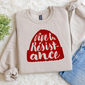 May include: A beige sweatshirt with a red beanie graphic. The beanie has the words "Vive la Résistance" in white script. The sweatshirt is made of a soft material and is suitable for casual wear.