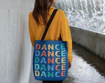 以下が含まれることがあります： 虹色の「DANCE」という文字が5回繰り返された青いトートバッグ。長い茶色の髪と黄色のセーターを着た人が持っています。