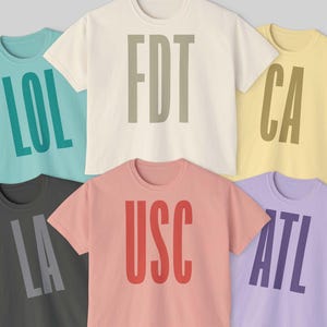 May include: A collection of six short-sleeved t-shirts in various colors, each displaying bold, capitalized text. The shirts are in shades of teal, cream, yellow, gray, coral, and lavender. The text on the shirts reads "LOL", "FDT", "CA", "LA", "USC", and "ATL".