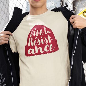 May include: Cream-colored t-shirt featuring a red graphic of a beanie with the words "Vive la Résistance" in white script. The shirt is made of a soft material and is suitable for casual wear.