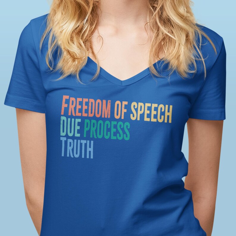 May include: A blue V-neck t-shirt featuring the words "FREEDOM OF SPEECH DUE PROCESS TRUTH" in orange, yellow, and green. The shirt is a solid blue colour and has a simple, classic design.