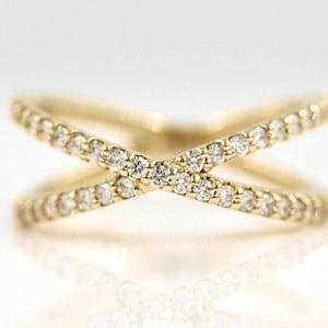 Könnte beinhalten: Ein goldfarbener Ring mit Kreuzmuster, besetzt mit kleinen, klaren Edelsteinen. Der Ring ist vor einem weißen Hintergrund platziert, mit einer darunter sichtbaren Reflexion. Das Design des Rings besteht aus zwei Bändern, die sich kreuzen und eine X-Form bilden.