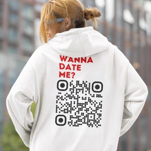 May include: A white hooded sweatshirt featuring the red text "WANNA DATE ME?" and a large black QR code. The garment is worn by a person with blonde hair.