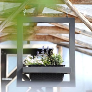 Puede incluir: Una jardinera colgante de metal gris oscuro con un marco rectangular. La jardinera contiene una variedad de plantas con flores blancas y moradas y follaje verde. La jardinera está suspendida de una estructura de madera.