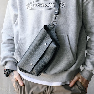 May include: A black leather crossbody bag with a geometric pattern and a long strap. The bag is worn over a gray hoodie with a white logo and olive green beanie. The person is wearing brown pants.