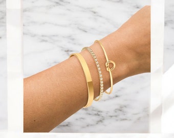 May include: Three gold bracelets on a wrist. The bracelets are a thin gold bangle, a thicker gold bangle, and a gold bangle with small clear stones.