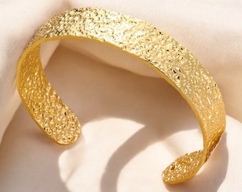 Könnte beinhalten: Ein goldfarbenes Armreif-Armband mit strukturierter, gehammerter Oberfläche. Das offene Design ermöglicht ein einfaches Tragen. Das Schmuckstück wird auf einem cremefarbenen Stoff präsentiert.