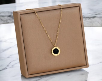 May include: A gold necklace with a round pendant featuring black Roman numerals on a gold background. The necklace is displayed in a brown velvet box.