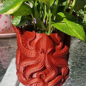 May include: A red octopus-themed planter pot with a detailed design. The pot is filled with green and yellow leafy plants. The octopus design features tentacles and a detailed face. The pot is made of a solid material.