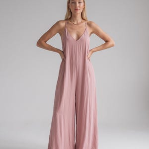 Könnte beinhalten: Ein altrosa Jumpsuit mit tiefem V-Ausschnitt, dünnen Spaghettiträgern, die sich am Rücken kreuzen, und einer weit geschnittenen Hose. Das Kleidungsstück besteht aus einem leichten, strukturierten Stoff, der in einer fließenden Silhouette fällt.