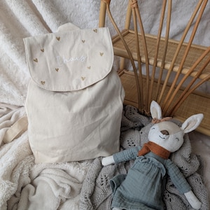 Può includere: Zaino in tela beige con patta, decorato con cuori dorati e il nome "Thais" in corsivo. Una bambola a forma di cervo con un vestito blu e una sciarpa arancione è appoggiata accanto.