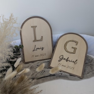 Könnte beinhalten: Zwei Holzschilder mit gewölbten Oberteilen. Jedes Schild hat einen dunkelbraunen Rand und eine helle Holzmitte. Eines zeigt den Buchstaben "L" und den Namen "Louiz" mit dem Datum "11 mai 2025". Das andere hat den Buchstaben "G" und den Namen "Gabriel" mit demselben Datum.