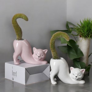 Op de afbeelding: Twee keramische plantenbakken in de vorm van een kat met een glanzende roze en witte afwerking. Elke kat staat in een uitgerekte houding met een gebogen groene cactus op de rug. De roze kat staat op een witte doos met gedrukte tekst.
