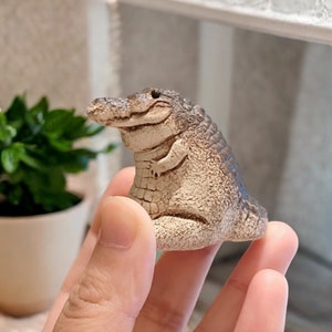 May include: A small, textured, beige and brown figurine of a seated alligator with a detailed scaly back, a wide snout, and a smooth underbelly. The creature is depicted in a sitting position with its front limbs resting against its body.