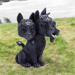 May include: A black three-headed dog figurine with large dark eyes, pointed ears, and a textured coat. Each head features a silver pentagram and crescent moon symbol on the forehead. The creature wears a studded collar with a small skull charm.