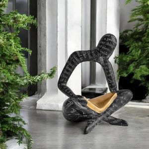 Könnte beinhalten: Eine schwarze Skulptur einer sitzenden Figur mit gekreuzten Beinen. Die Oberfläche ist mit weißem Text bedruckt. Die Figur weist im Schoßbereich einen glatten, goldfarbenen Innenbereich auf, der einen Kontrast zur dunklen Außenseite bildet.