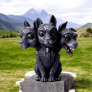 May include: A black three-headed cat statue with a pentagram on its forehead and a skull on its collar. The statue is sitting on a stone pedestal in a grassy field.