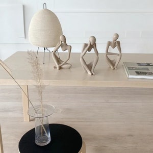 Pode incluir: Três esculturas humanas abstratas de cor bege em poses sentadas e contemplativas. Cada figura tem uma superfície lisa e texturizada e repousa sobre uma mesa de madeira clara ao lado de uma lâmpada branca, um livro e um vaso de vidro.