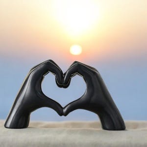 May include: A smooth, matte black sculpture depicting two hands positioned together to form a heart shape. The object is solid in color with defined fingers and knuckles, creating a symmetrical silhouette that frames the light in the background.