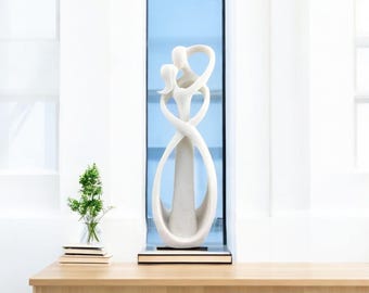May include: A tall, off-white abstract sculpture depicting two stylized figures intertwined in a continuous, flowing form. The smooth, matte-finished piece features curved lines that create an open, oval-shaped base, standing on a dark rectangular plinth.