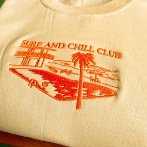 Può includere: Felpa color crema con un design ricamato arancione. Il design presenta una palma, una barca e il testo "SURF AND CHILL CLUB". La felpa è realizzata in un materiale morbido.