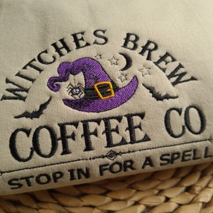 May include: A beige sweatshirt with an embroidered design that says "Witches Brew Coffee Co." and "Stop in for a spell". The design features a purple witch's hat with a spider and a yellow buckle.