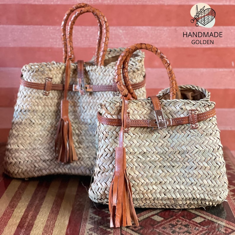 Puede incluir: Dos bolsos de paja trenzada con detalles de cuero marr&oacute;n. Los bolsos tienen asas trenzadas, correas de cuero con hebillas y borlas. Los bolsos est&aacute;n etiquetados como "HANDMADE GOLDEN".