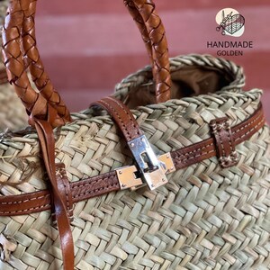 Puede incluir: Bolso tote de paja tejida con detalles de cuero marr&oacute;n. El bolso tiene un asa de cuero trenzado y un cierre de hebilla plateada. El texto "HANDMADE GOLDEN" es visible.
