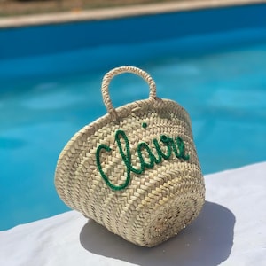 Peut inclure: Un sac cabas en paille tressée avec le nom "Claire" brodé en vert sur le devant. Le sac est posé sur une surface blanche près d'une piscine bleue.