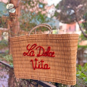May include: Woven rectangular tote bag with two handles. The bag is tan with the words "La Dolce Vita" embroidered in red. The bag has a rectangular shape and is made of woven material.