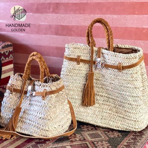 May include: Two woven straw handbags with brown leather accents. The bags have braided handles, leather straps, and decorative tassels. The smaller bag has a shoulder strap. The bags are labeled "HANDMADE GOLDEN".