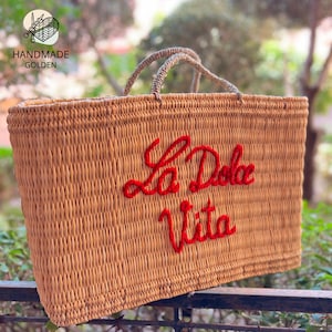 May include: A rectangular woven straw tote bag with red embroidered text that reads "La Dolce Vita". The bag has two rope handles and a woven texture. The bag is tan in color.