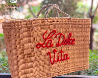 Puede incluir: Un bolso tote rectangular de paja tejida con texto bordado en rojo "La Dolce Vita". El bolso tiene dos asas de cuerda y una textura tejida. El bolso es de color marrón claro.