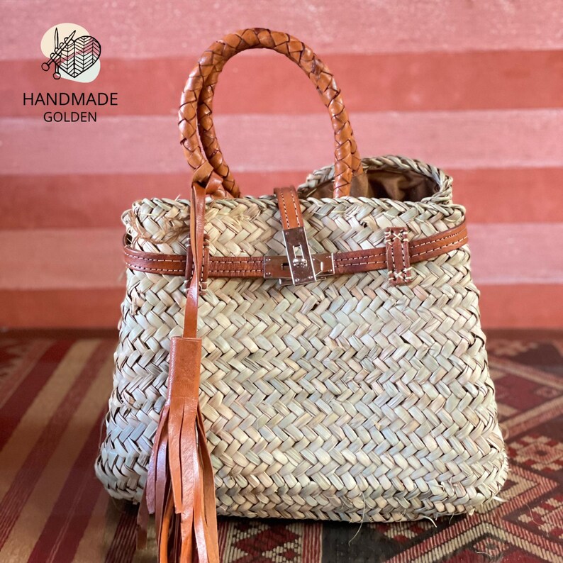 Puede incluir: Bolso de mano artesanal de paja tejida con asa trenzada de cuero marr&oacute;n y cierre de correa de cuero marr&oacute;n. El bolso presenta una borla de cuero marr&oacute;n y el texto "HANDMADE GOLDEN".