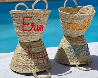 Puede incluir: Dos bolsos de mano de paja tejida con asas arqueadas. Una bolsa tiene el nombre "Erin" bordado en rojo, y la otra "Ashli" en amarillo. Debajo, los nombres "Happy" e "Yaya" están bordados en rosa y azul.