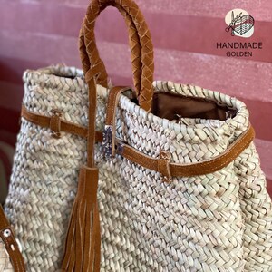 May include: Woven straw tote bag with brown leather accents. Features a braided handle, leather straps, and a decorative tassel. The bag has a metal clasp and the text "HANDMADE GOLDEN".