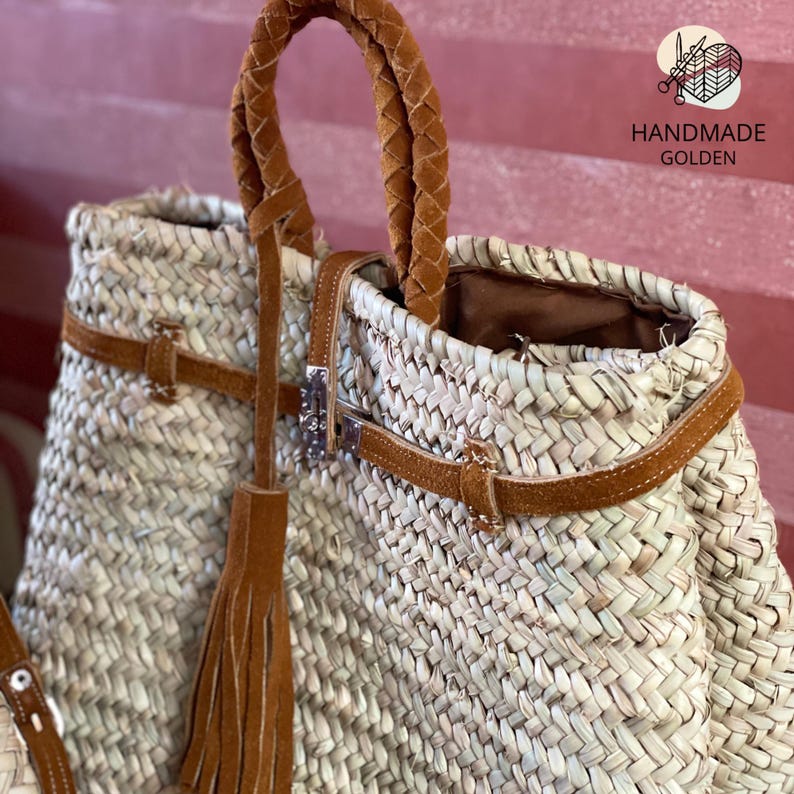 May include: Woven straw tote bag with brown leather accents. Features a braided handle, leather straps, and a decorative tassel. The bag has a metal clasp and the text "HANDMADE GOLDEN".